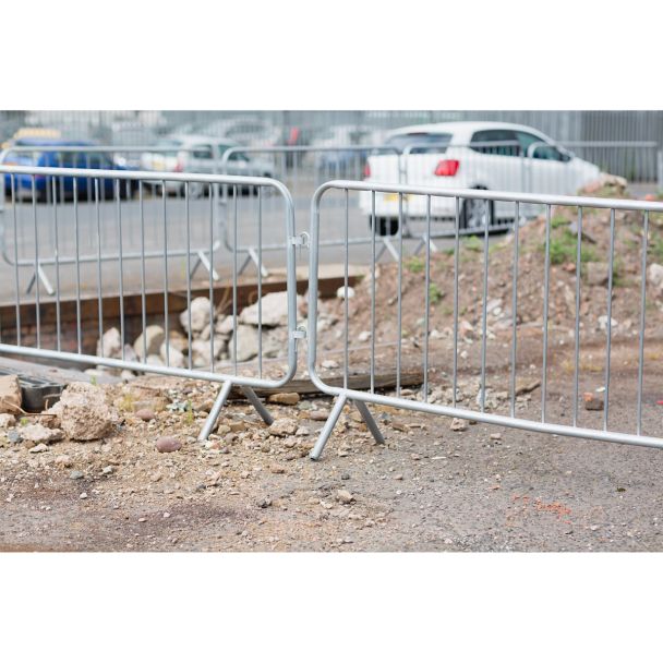 Multiple galvanised steel crowd barriers deployed on a gravel construction site, enclosing a hole in the ground.